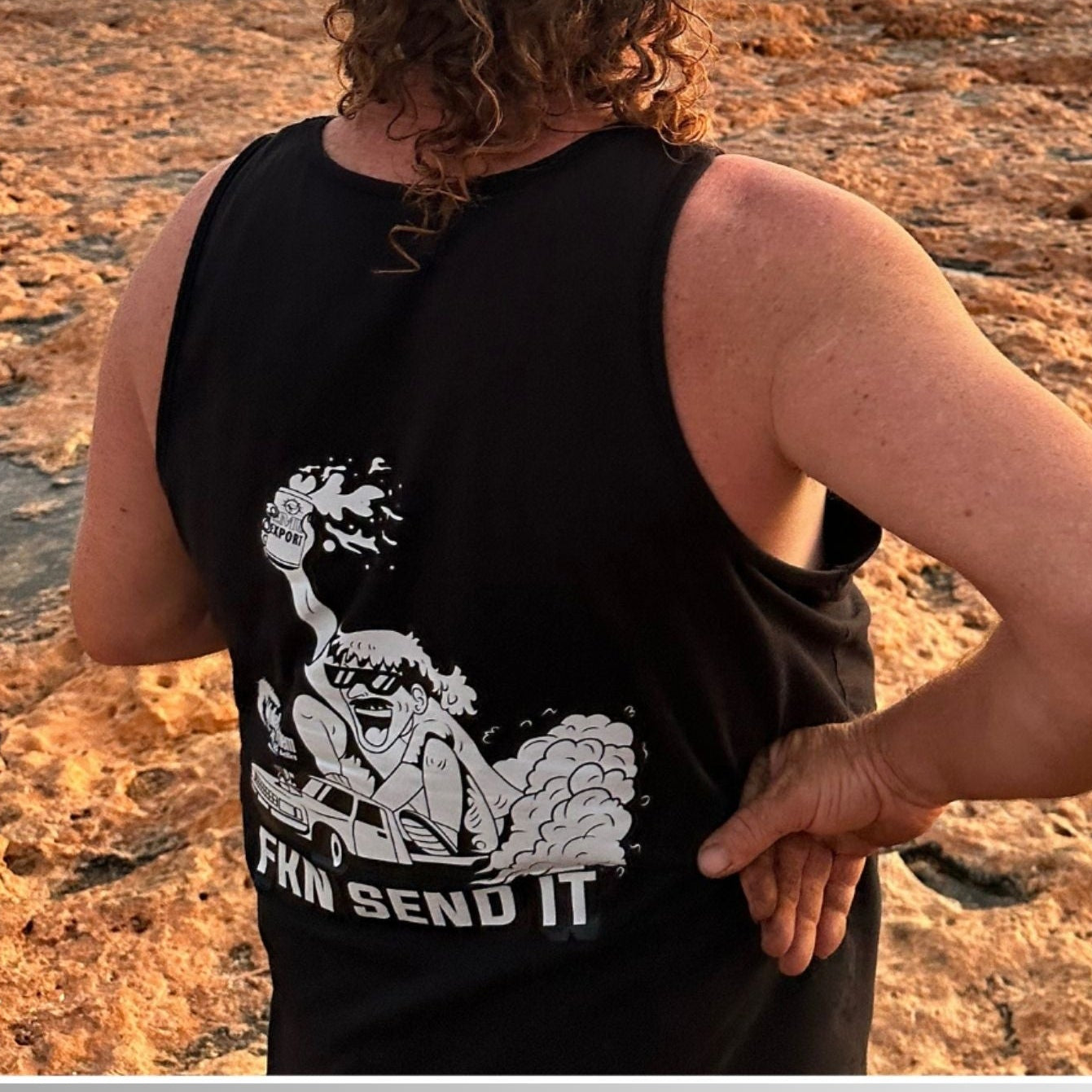Person wearing a black tank top with a graphic design, standing on a rocky beach at sunset.