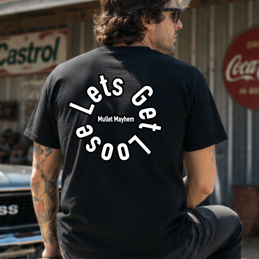 A man wearing a black t-shirt with a white graphic text design on the back, sitting in front of a rustic metal building with vintage Coca-Cola and Castrol signs.