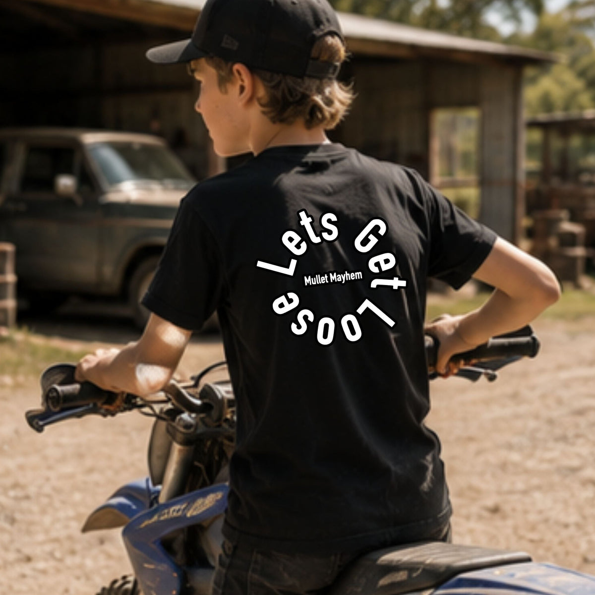 A boy wearing a black t-shirt with a white graphic on the back and a black cap, sitting on a blue dirt bike in an outdoor rural setting with a barn and truck in the background.