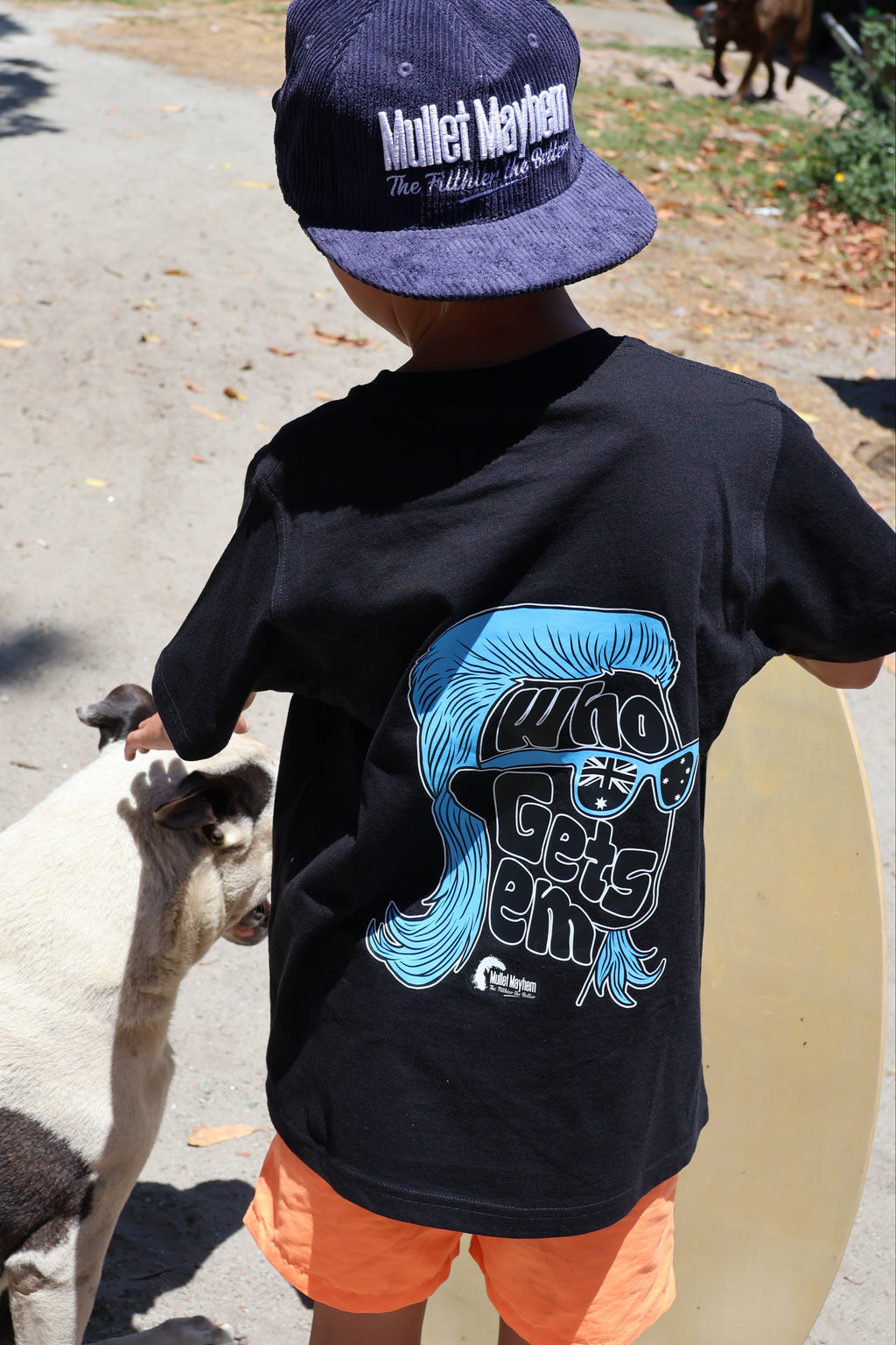 A child seen from behind wearing a black t-shirt with a blue and white graphic print, orange shorts, and a navy blue corduroy cap. The child is standing outdoors on a paved path next to a goat.
